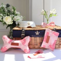 Pet hamper, featuring a Buckingham Palace wicker hemper, ceramic pet bowl with Windsor crest, pink bow tie, pet toy, bag and bandana bathed in sunshine with flowers decorating. 