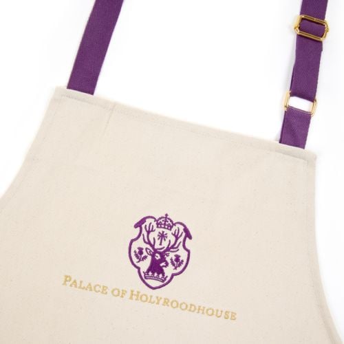 Cream coloured apron with deep purple adjustable straps and large central pocket, purple emblem featuring a stag with Palace of Holyroodhouse spelled out in golden thread at the top centre. 