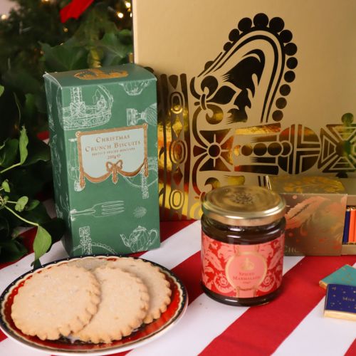 Buckingham Palace Gold Gift Box with a selection of festive treats set against a Christmas tree on a red and white striped tablecloth. 