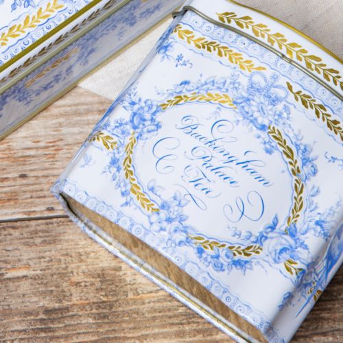 Close up shot of a white tea caddy with a floral blue design and the words 'Buckingham Palace Tea' in the centre of the tin. Surrounding the design is a leaf garland design in gold.