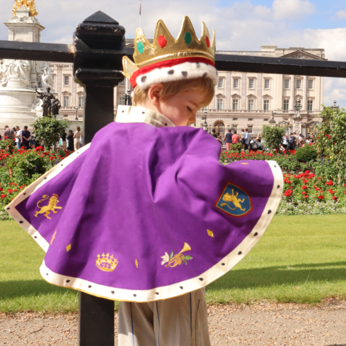 Child wearing cape and gold crown outside of Buckingham Palace