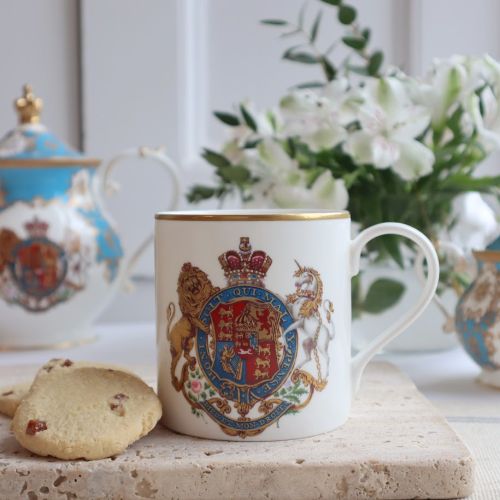 A white mug with royal crest, and handle with flowers in the background, biscuits in the foreground  and the rest of the Coat of Arms china selection. 