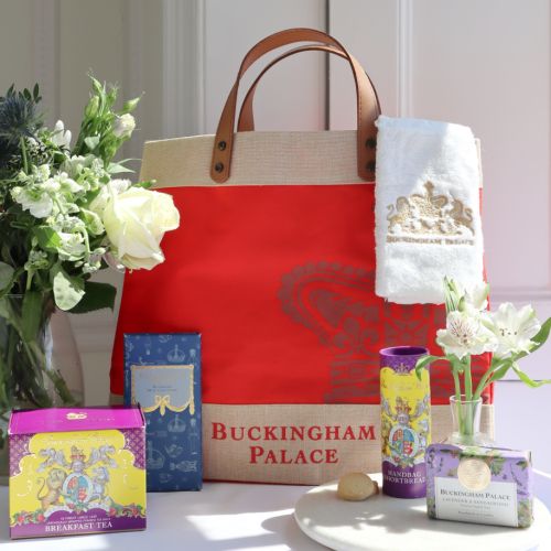 Buckingham Palace Mum to Be Gift Bag, featuring a red Buckingham Palace shopper bag, English Breakfast Tea, Shortbread Biscuits, Milk Chocolate bar, Buckingham Palace face towel and Soapsmith soap, with flowers in the background. 