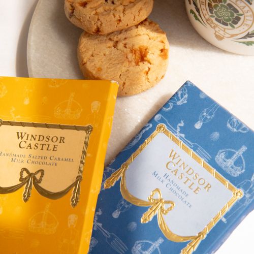 The Taste of Windsor Castle Hamper featuring a wicker hamper with blue lining and a crown as well as "Buckingham Palace" printed on the hamper. Items featured in the hamper include, Windsor Castle Fudge box with fudge in an adjacent marble bowl. Also feat