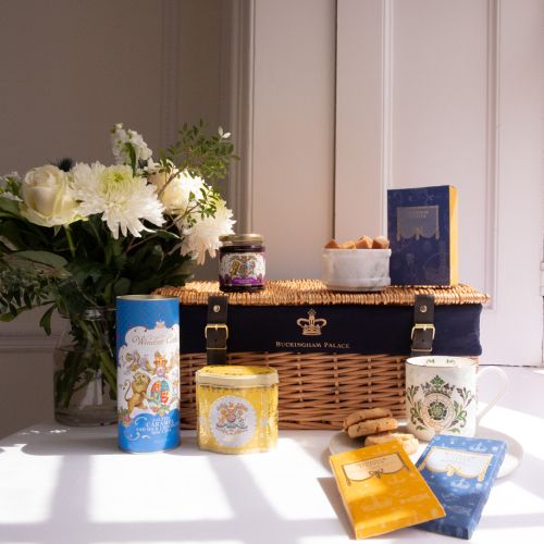 The Taste of Windsor Castle Hamper featuring a wicker hamper with blue lining and a crown as well as "Buckingham Palace" printed on the hamper. Items featured in the hamper include, Windsor Castle Fudge box with fudge in an adjacent marble bowl. Also feat