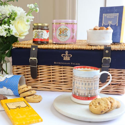 The Taste of Buckingham Palace Hamper featuring a wicker hamper with blue lining and a crown as well as "Buckingham Palace" printed on the hamper. Items featured in the hamper include, Windsor Castle Fudge box with fudge in an adjacent marble bowl, pink E