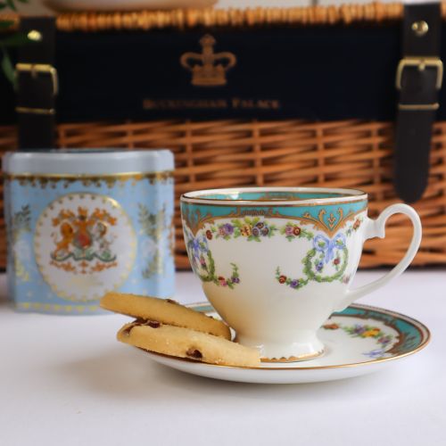 A wicker hamper with two Great Exhibition tea cups, a pink biscuit tin and a blue Earl Grey Tea Caddy. There are flowers to the left of this image. 