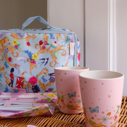 The Picnic on the Palace Lawn, featuring the Summertime Picnic Range with a blue patterned lunch bag and plates with Royal Guards, butterflies, birds  and Guardsman and a pink pattern, on a cutlery wrap and cups featuring a royal crown and floral pattern.