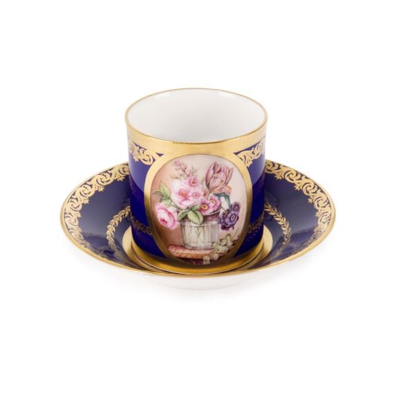 The Prestige Vase of Flowers Gobelet Litron, featuring central oval shape with hand painted bouquet of pink and purple flowers in a wooden pot. The rest of the teacup and saucer has a rich cobalt blue paint and is finished with 22 carat gold gilding along