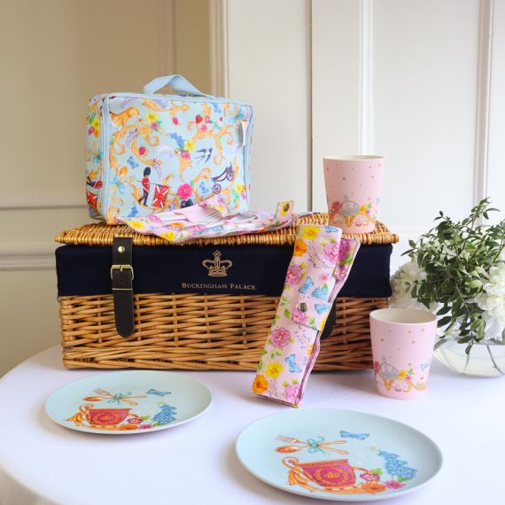 The Picnic on the Palace Lawn, featuring the Summertime Picnic Range with a blue patterned lunch bag and plates with Royal Guards, butterflies, birds  and Guardsman and a pink pattern, on a cutlery wrap and cups featuring a royal crown and floral pattern.