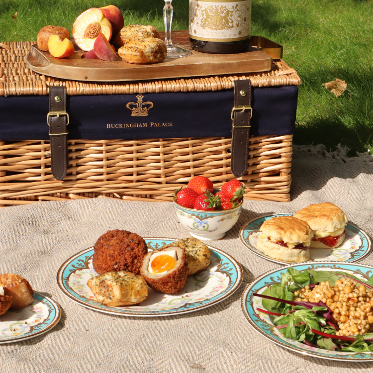 A picnic basket on a blanket with a variety of food on small plates including Scotch Eggs and Sausage Rolls